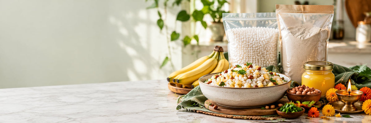 A bowl of sabudana khichdi with bananas, peanuts, a jar of ghee, bags of sabudana and farali flour, marigolds and a lit diya on a marble kitchen table